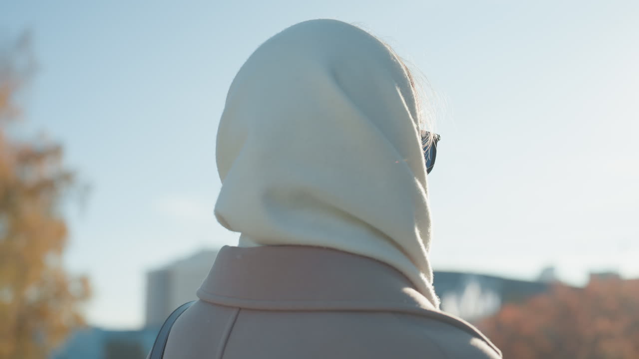 Donna con foulard retroilluminato dal sole, in piedi su un viale cittadino. La sua silhouette è ammorbidita da bagliore e alone. Alberi autunnali ed edifici distanti. Indossa un cappotto beige e occhiali da sole. Umore riflessivo e speranzoso, una pendolare.