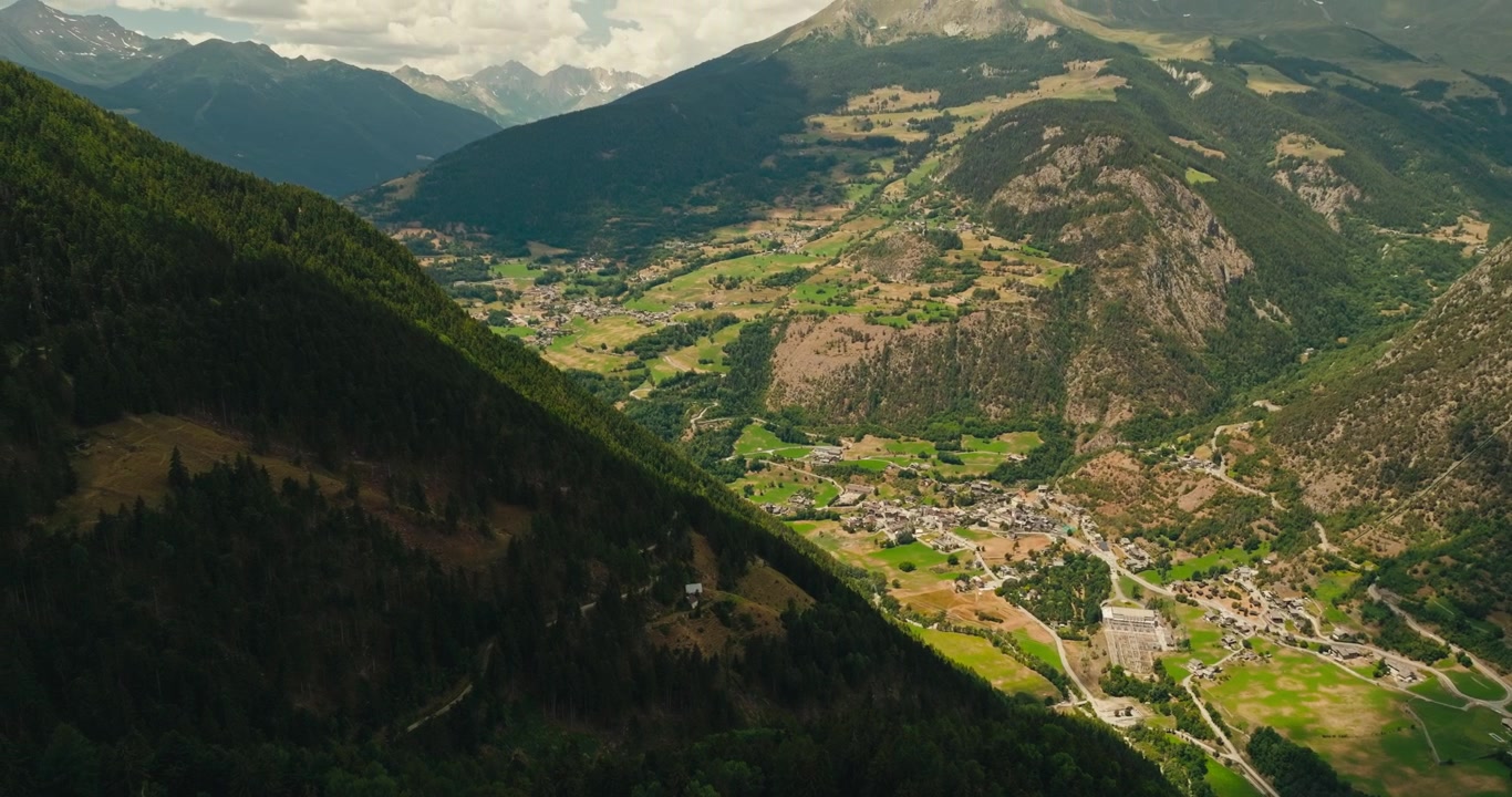 Drone vola in alto sopra un villaggio di montagna che domina una vasta valle alpina con lontane cime montuose sotto un drammatico cielo estivo nuvoloso, catturando paesaggi mozzafiato