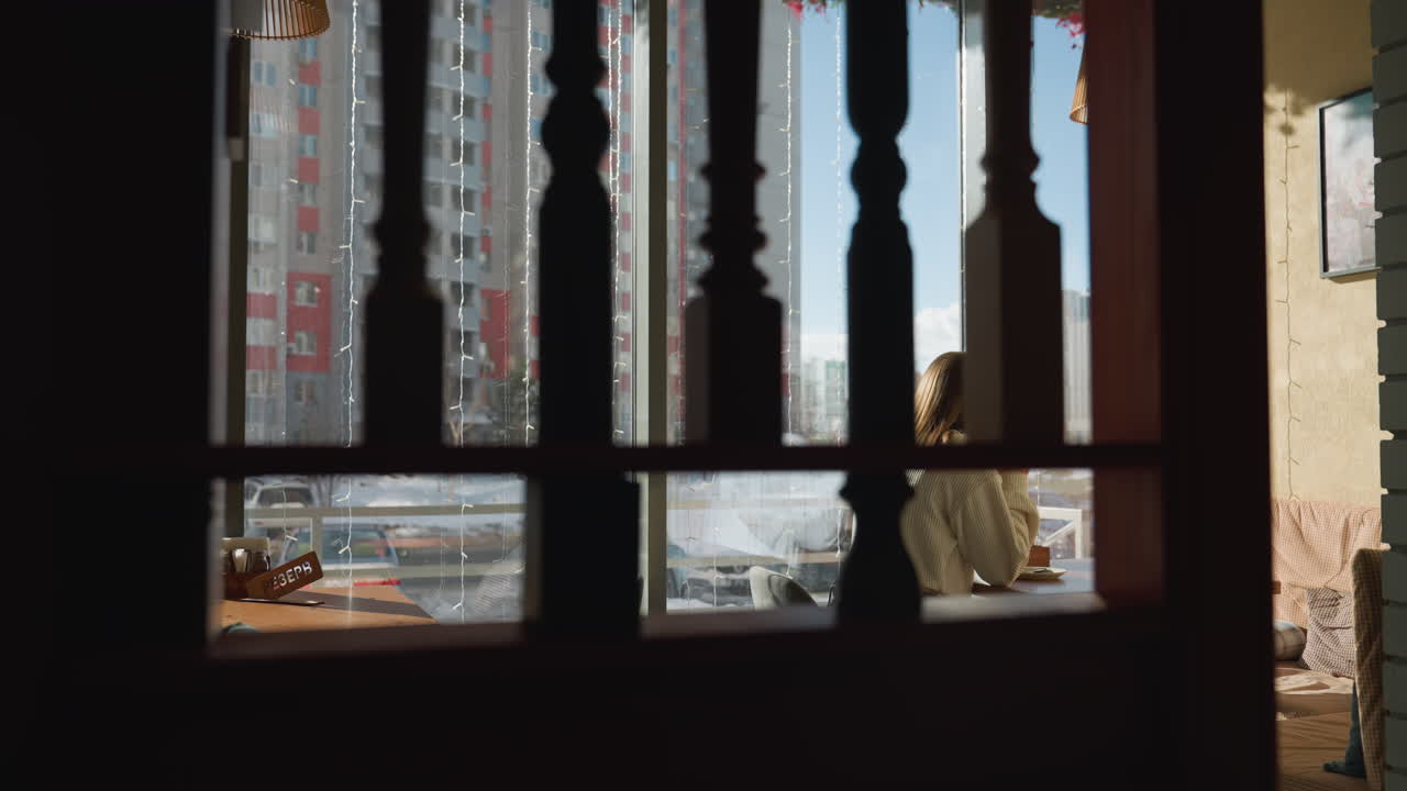 View through wooden frame shows artist seated at window-side table sipping tea, soft winter sunlight pouring in through glass panels, city buildings and parked cars visible in calm urban background