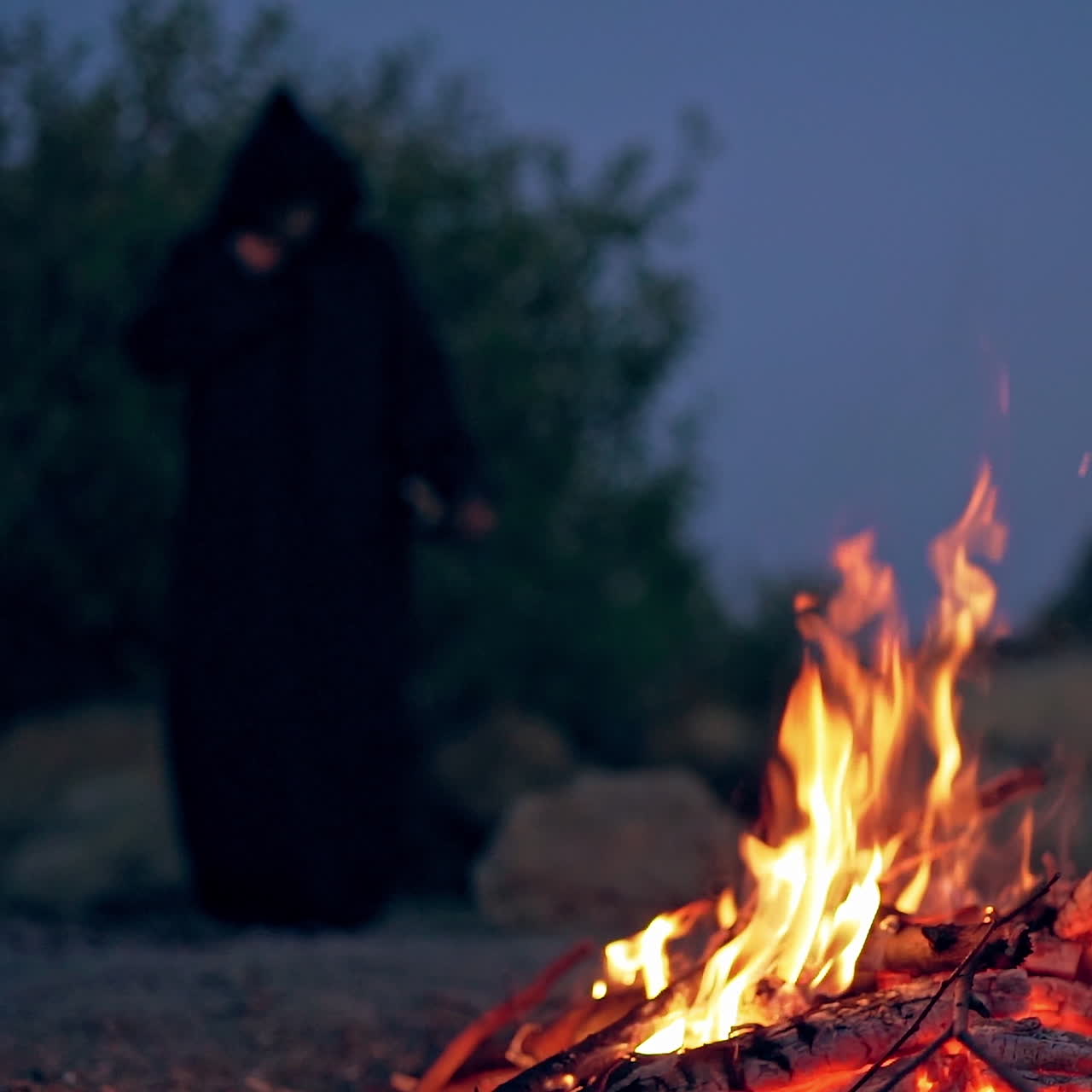 Halloween. Burning up wood on the natural background with dark figure. Scary beldam standing behind the fire outdoors in the evening.