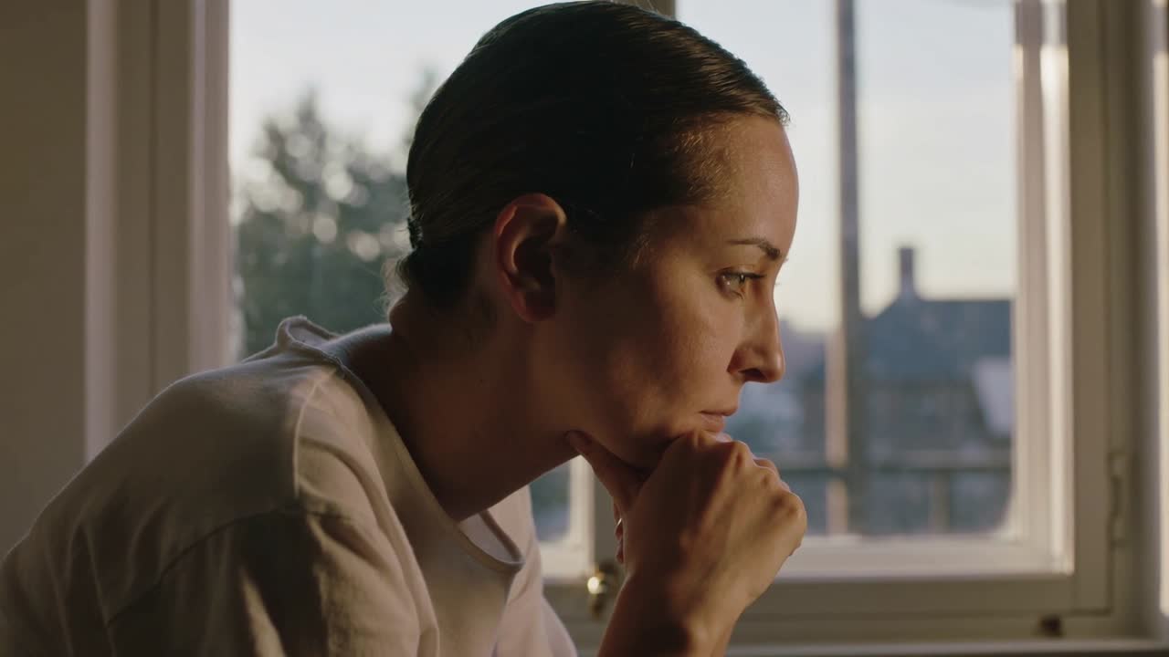 Gazing woman in light shirt reflecting while propping chin at multi-pane window, sunlight shifting