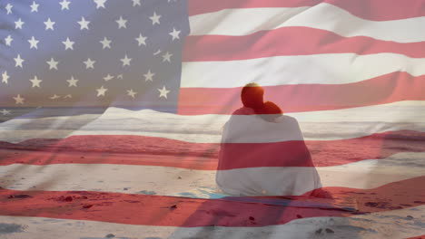 animation of american flag over happy diverse couple wrapped in rug embracing sat on sunset beach