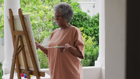 Ältere-Afroamerikanische-Frau-Malt,-Während-Sie-Auf-Der-Veranda-Des-Hauses-Steht