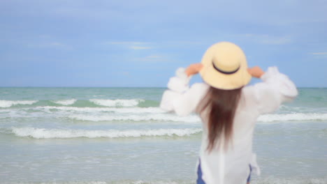 a young woman in a flowing white shirt, sun hat, and shorts as she jogs into the frame toward the incoming surf