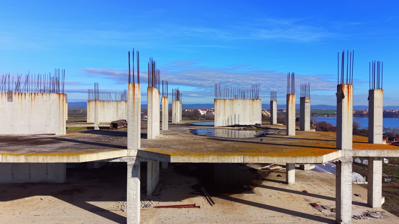 Rooftop view of building under development featuring concrete pillars with steel armature and water puddles. Top floor of concrete construction site with vertical rebar. Aerial view