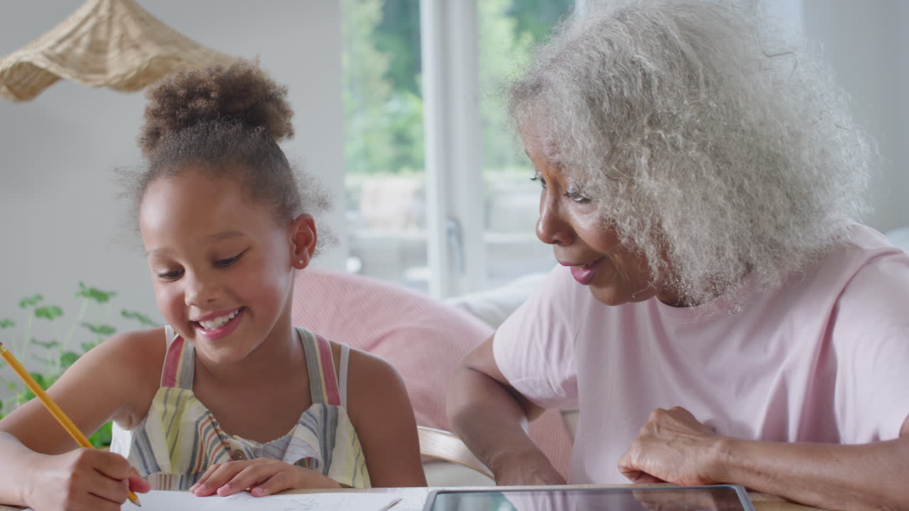 Grandmother Helping Granddaughter With Home Schooling Sitting At Table With Digital Tablet