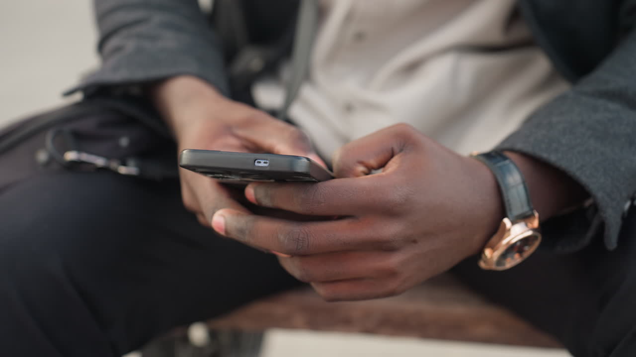 Mãos segurando um smartphone ao lado de uma bolsa de notebook colocada perto das coxas, destacando o estilo moderno e casual, com foco no relógio de pulso, no gesto humano e na conexão tecnológica expressa por meio do equilíbrio