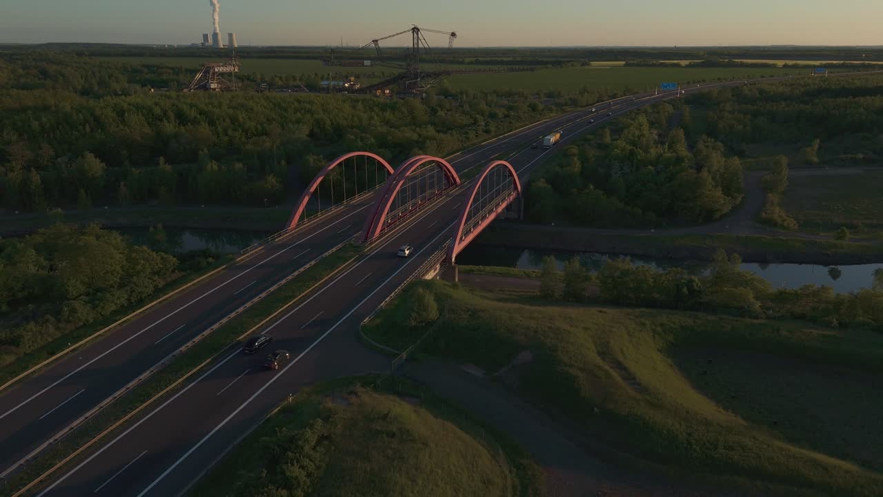 Wide drone shot near Leipzig showing a highway bridge, expansive landscape, a massive coal excavator, and a distant power plant. Captures industrial infrastructure in evening light.