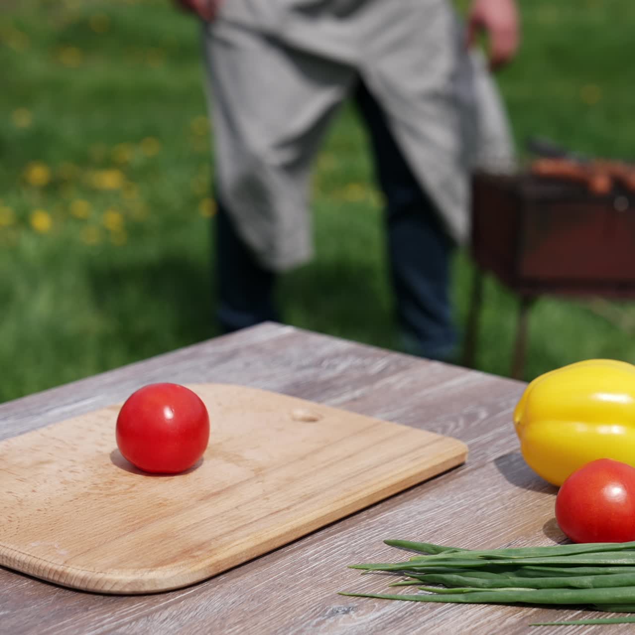 Fresh vegetables on wooden table outdoors. Food for picnic on the blur background of nature. Man frying sausages on a brazier