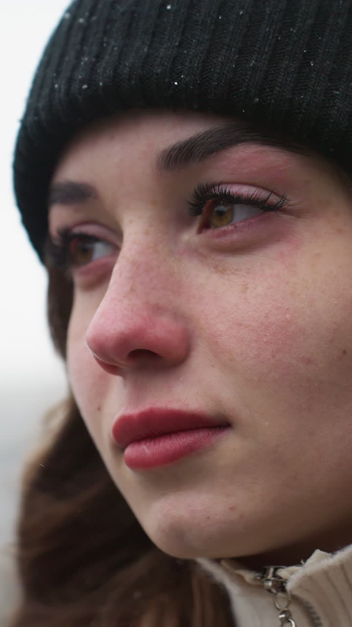 Close up of thoughtful girl in black cap and brown jacket walking with serious expression and wind-blown hair against blurred city background during cold quiet day with overcast sky