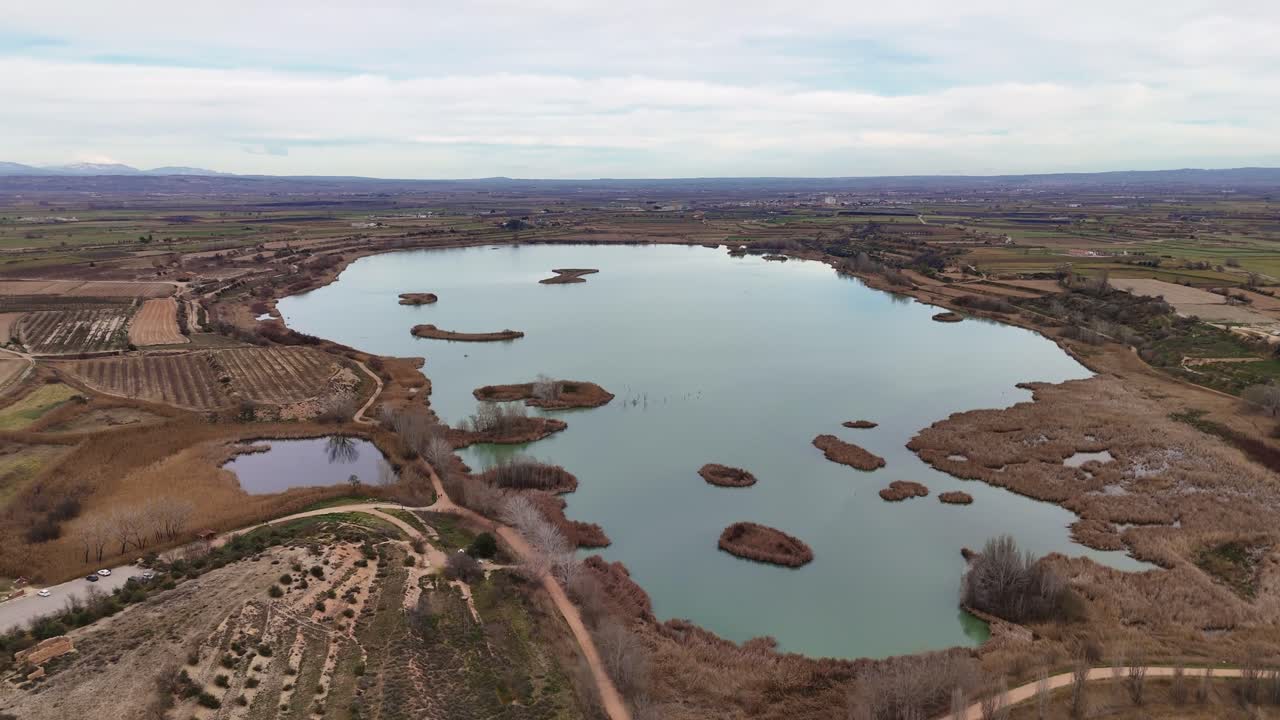 Aerial view of a vast lake with patches of marshland and small islands, surrounded by farmland and sparse vegetation under a cloudy sky