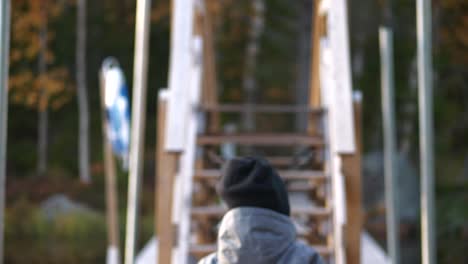 Dolly-shot-of-Young-boy-walking-over-a-bridge-on-a-hiking-trail-surrounded-by-autumn-colors,-pretending-to-hold-weapons-in-his-hands