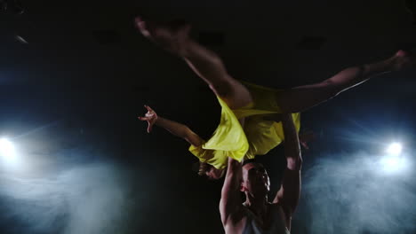 two dancers a man and a woman run to each other and a male partner raises a woman in a yellow dress in her arms and rotates in the air performing top support.