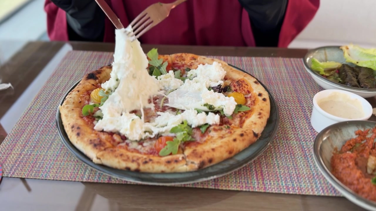 Primo piano di mani che usano coltello e forchetta per tagliare una fresca pizza con burrata, pomodori e rucola. Una persona si gode un pranzo diurno a un tavolo di ristorante con contorni.