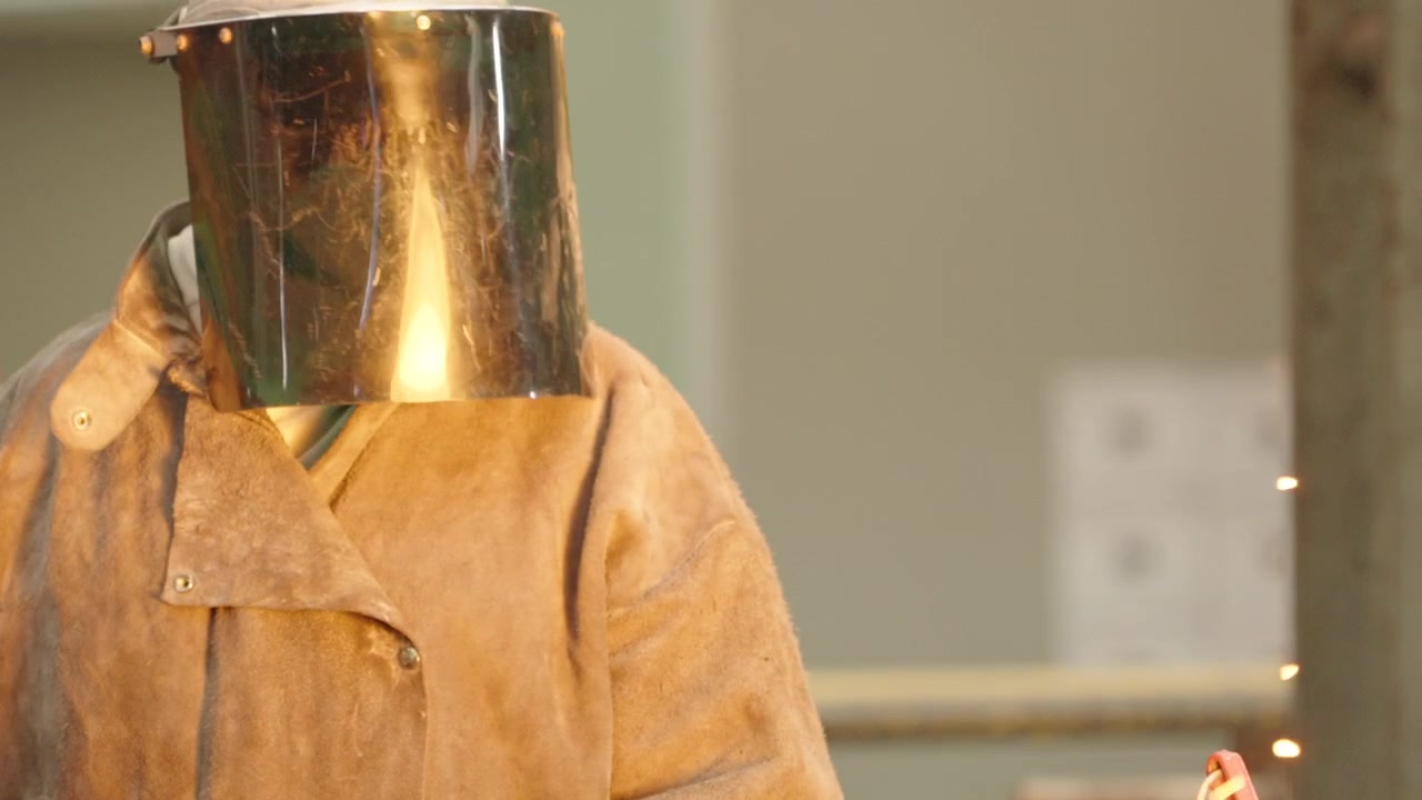Operator in a heat resistant coat and reflective visor stands still inside a bright engineering steel foundry.