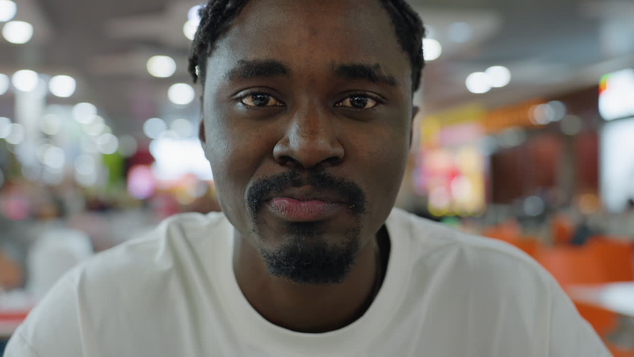 Close up of young man in white shirt biting burger with eyes closed while savoring taste, sitting inside colorful food court with blurred background lights and vibrant atmosphere