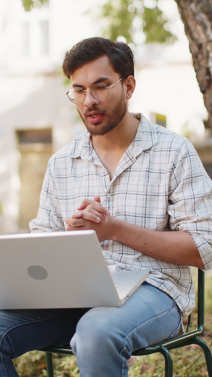 joven indio independiente sentado en una silla mirando la cámara del portátil haciendo una llamada de video al aire libre