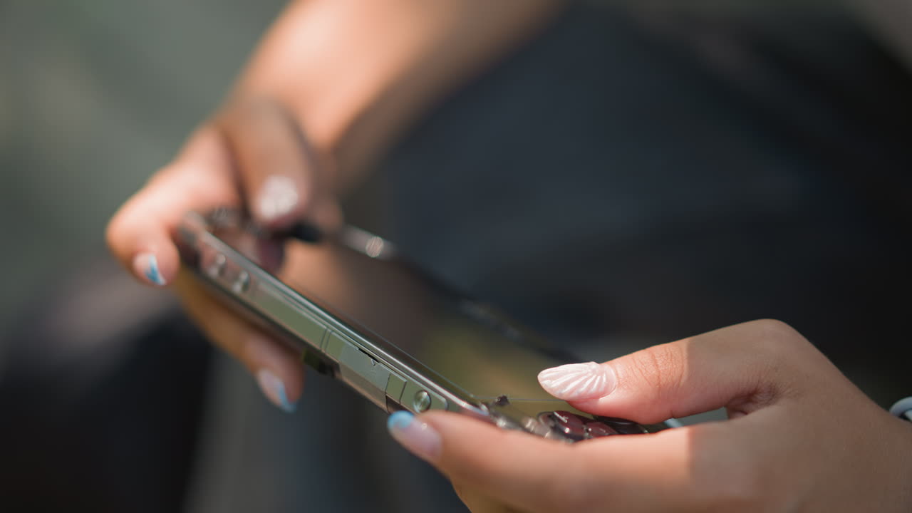 primer plano de unas manos sujetando un smartphone en el exterior, con uñas cuidadas que tocan la pantalla táctil, una elegante funda que refleja la luz del sol, poca profundidad de campo, desplazamiento casual por las apps, ambiente urbano relajado, influencer