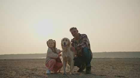 retrato de un feliz hombre de mediana edad con cabello gris en una camisa a cuadros que se sienta cerca de su gran perro de color crema y su pequeña hija rubia en una chaqueta blanca y pantalones rosados en una orilla desierta durante sus vacaciones fuera de la ciudad en una noche de verano