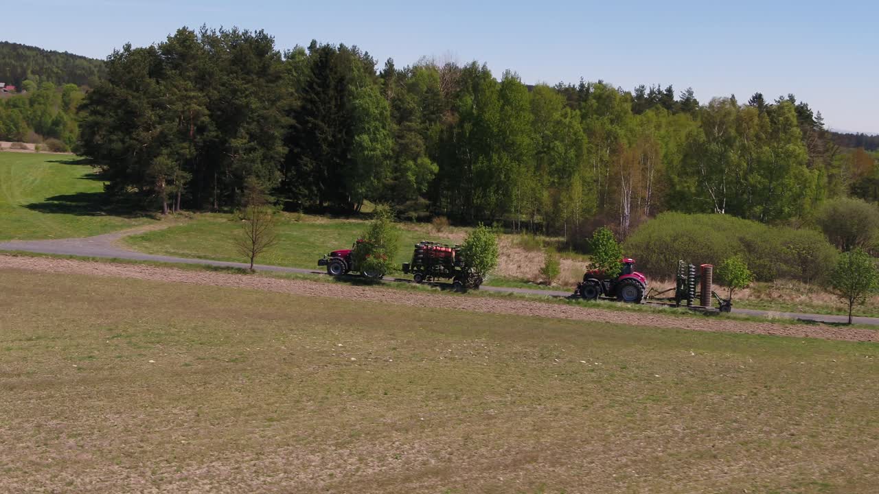 Drone semi orbit showing agricultural machinery resting beside field and tree line on a sunny spring day