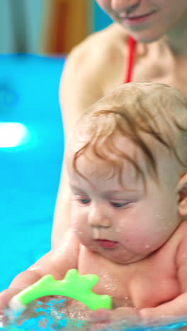 adorable caucasian baby with blond hair is learning to swim. coach moves the kid in the water while child is focused on toy. vertical video