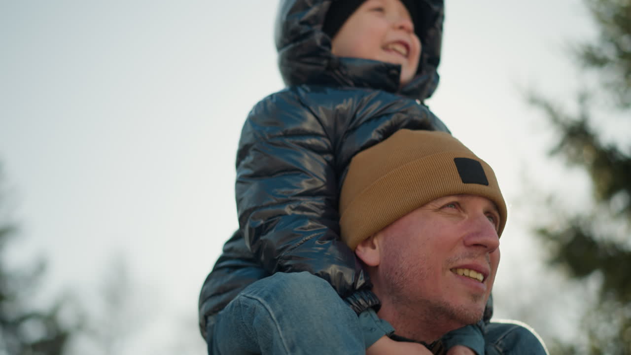 Close-up of a father carrying his joyful son on his shoulders as the boy nods his head happily, with warm sunlight shining brightly over them