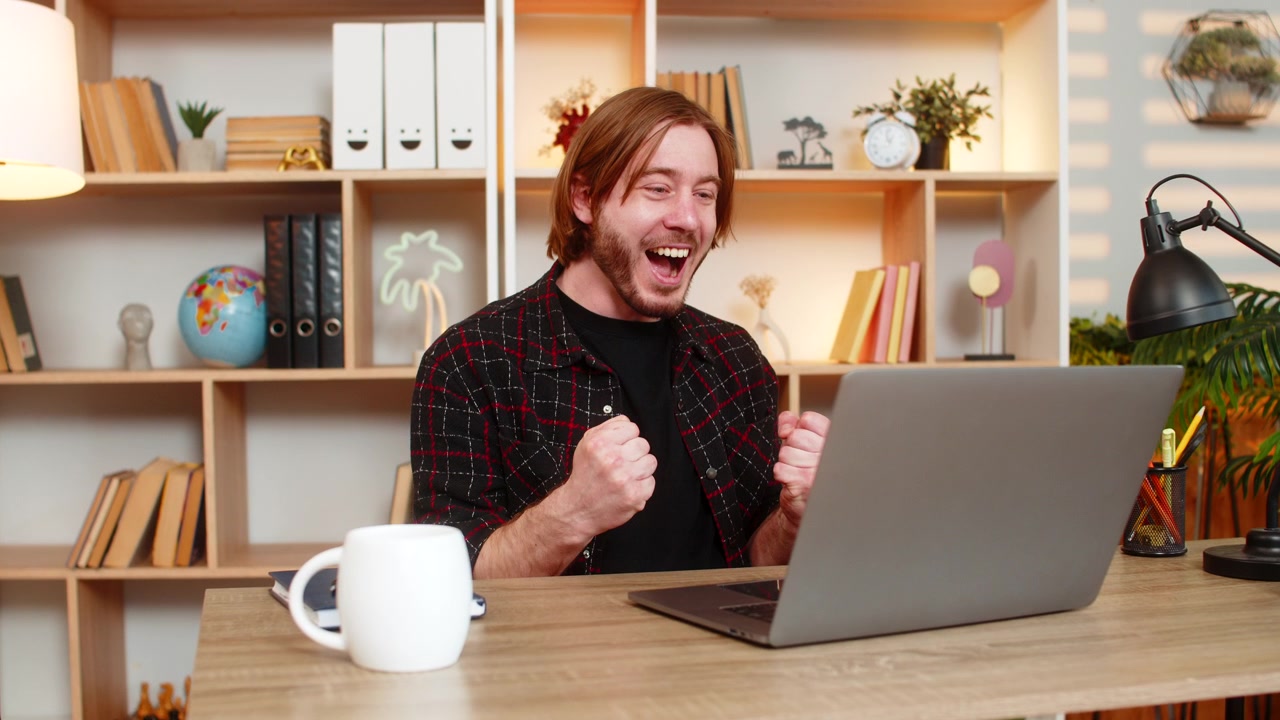 Giovane uomo al tavolo di casa che festeggia il successo con il laptop dopo aver visto un ottimo risultato online sullo schermo