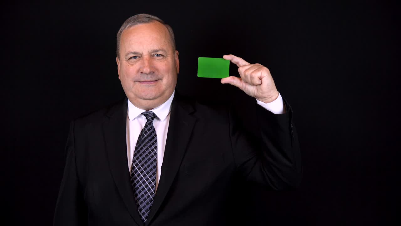 Old businessman in a suit holds a bank card in his hand. Chromakey green card. Isolated black background.