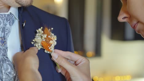 woman gently pins a rustic boutonniere to a man's dark suit with a patterned cravat