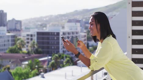 mixed race gender fluid person drinking cup of coffee and using smartphone on roof terrace
