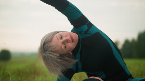 woman lying on one side, practicing side bend yoga pose with arm extended in a vast grassy field under cloudy skies, with trees blurred in the distance and a hint of something blurred nearby