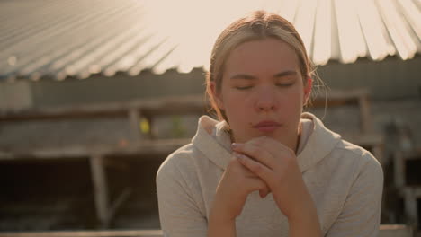 close-up of contemplative woman sitting on rustic stadium bleachers, hands locked together near her face, she appears reflective, with a soft expression against a blurred background