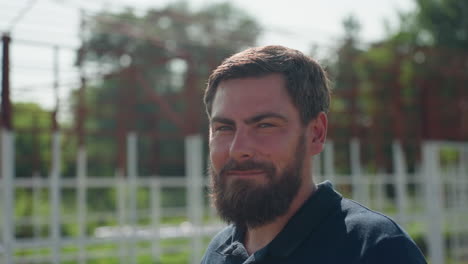 man turns toward camera with slight smile near metal farm frame and green plants, side profile showing beard, outdoor rural setting with blurred structure and trees in background