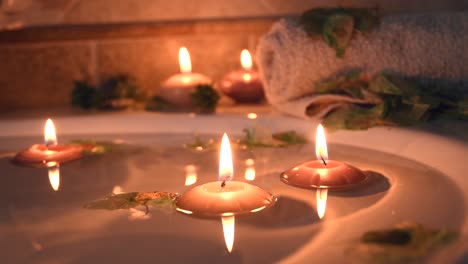 relaxing-spa-background-with-candles-floating-in-the-bath-water,-some-green-petals-and-a-towel-near-the-water-surface