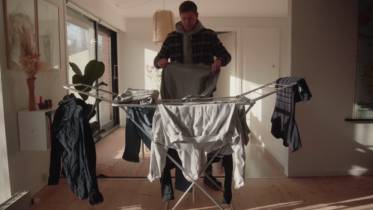 Young Male collecting and folding dry clean Clothes from Dryer At Home in sunlit modern living room 4K