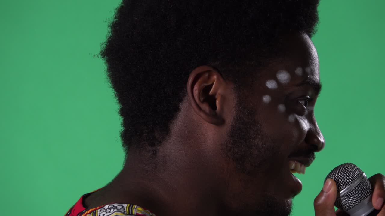마이크로폰으로 노래를 부르는 젊은 아프리카계 미국인 남성의 초상화: 얼굴에 민족적 백인 패턴을 가진 흑인 남성이 스튜디오에서 녹색 화면에 포즈를 취하고 있습니다.