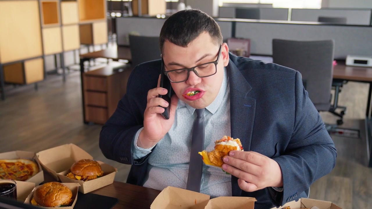 Overweight Businessman Eating Burger and Talking on Phone in Office