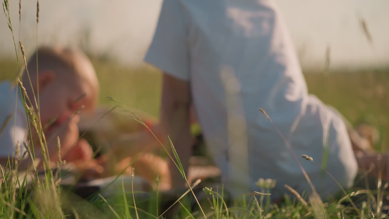 Blurry shot of two kids sitting on a checkered scarf in a grassy field, with a loaf of bread also in the frame. outdoor moment, emphasizing the simplicity of a countryside picnic