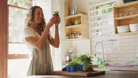 Smiling-caucasian-woman-tending-to-potted-plants-standing-in-sunny-cottage-kitchen