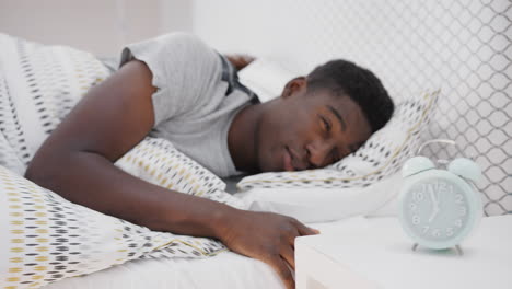 un hombre dormido en la cama extiende la mano para apagar el despertador en la mesa de noche.