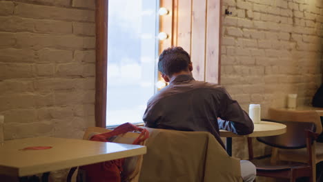 Man in formal shirt sitting alone at cozy cafe table near window, seen from behind, writing or working in calm ambient setting with warm lights, showcasing quiet concentration and creative focus