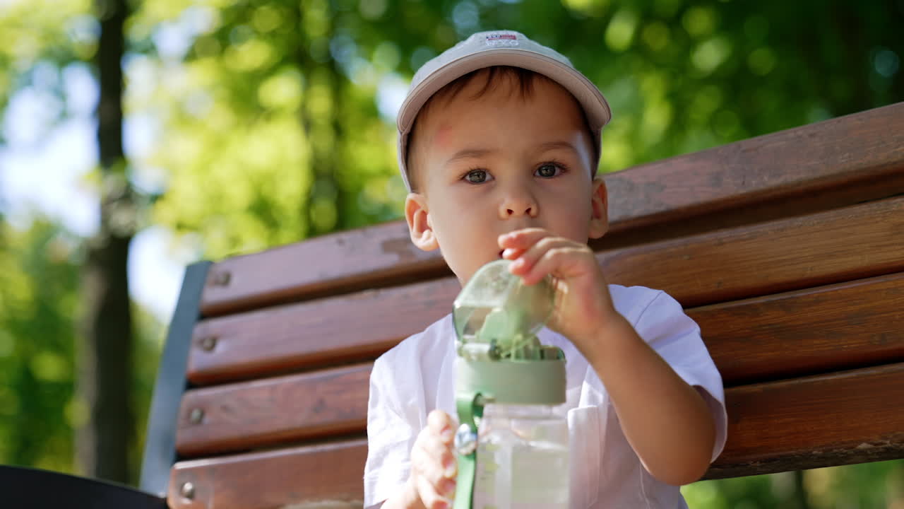 Caucasian baby boy in a cap sits on the bench holding sport bottle. Toddler opens the bottle ready to drink. Low angle view. Blurred nature backdrop.