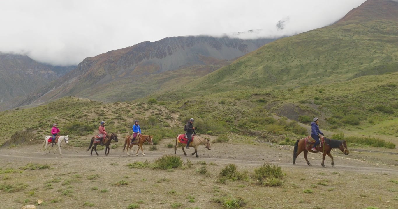 Uma tomada cinematográfica de drone com movimento de aproximação avança sobre cavaleiros viajando por terreno aberto, cercados por colinas verdes ondulantes e montanhas acidentadas do Himalaia em Mustang, Nepal.