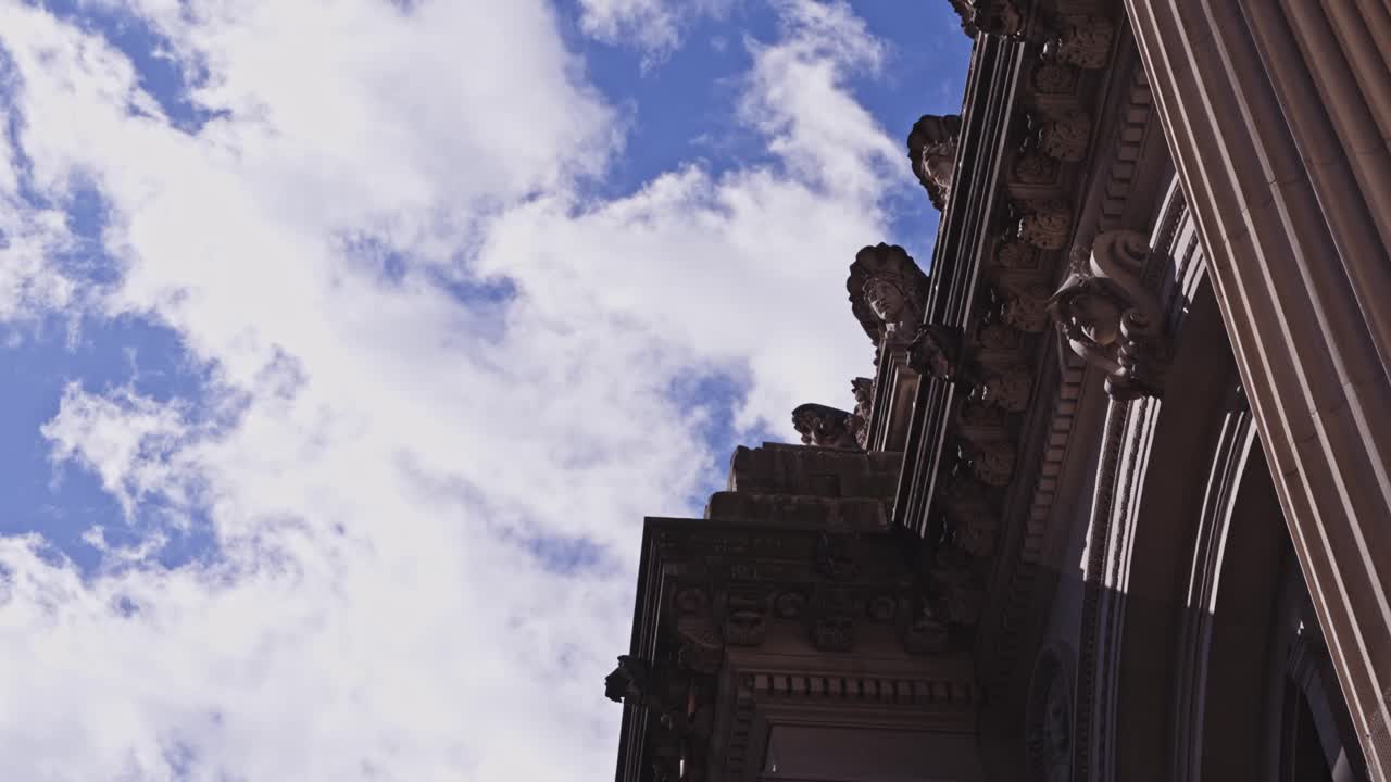 Dramatic upward angle of ornate stone carvings and Corinthian columns on a classical facade, framed by soft daylight clouds overhead