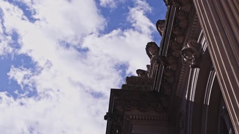 dramatic upward angle of ornate stone carvings and corinthian columns on a classical facade, framed by soft daylight clouds overhead