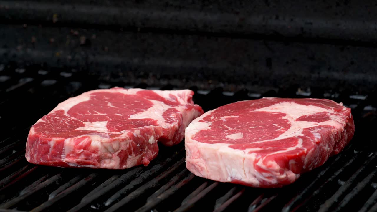 Two raw rib-eye steaks getting ready to cook on a grill