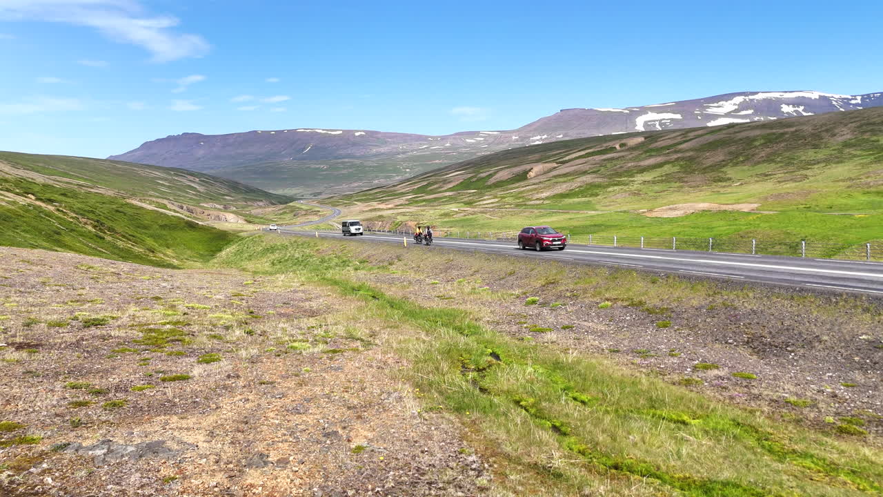 Flygvy med drönare över två cyklister som cyklar längs en slingrande väg genom en grön canyon med en flod på den isländska landsbygden, vilket symboliserar äventyr, frihet och hållbart resande