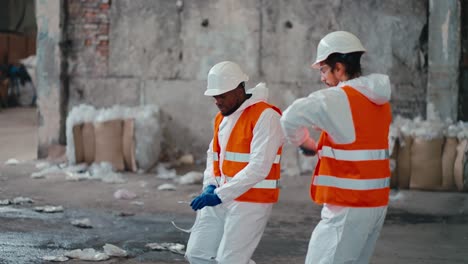 A-man-with-Black-skin-in-a-white-protective-uniform-and-an-orange-vest-together-with-his-colleague-a-man-with-a-beard-in-a-white-protective-helmet-dances-in-the-large-hall-of-a-waste-processing-and-sorting-plant