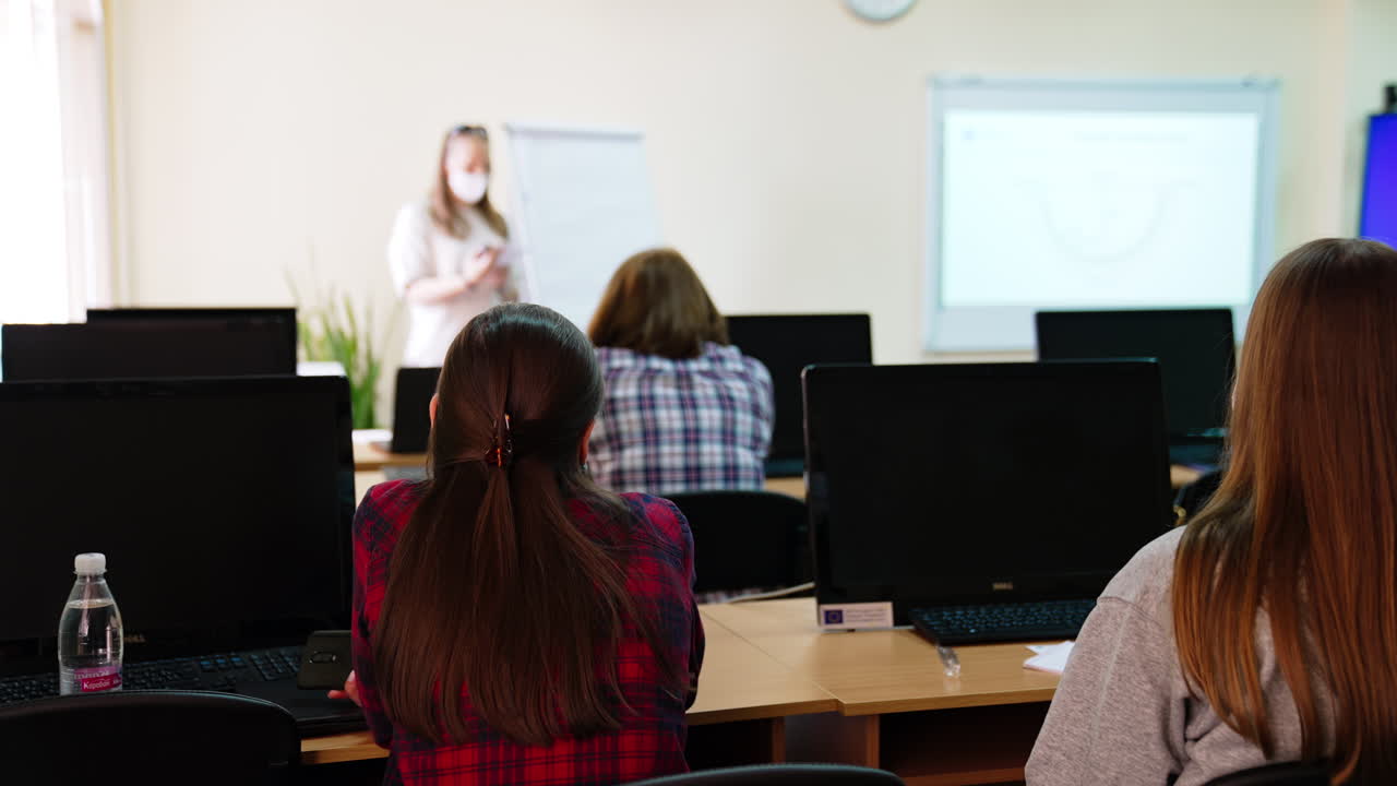 Rear view of the female students listening to the lecture. Lecturer at the whiteboard and screen showing images at backdrop.