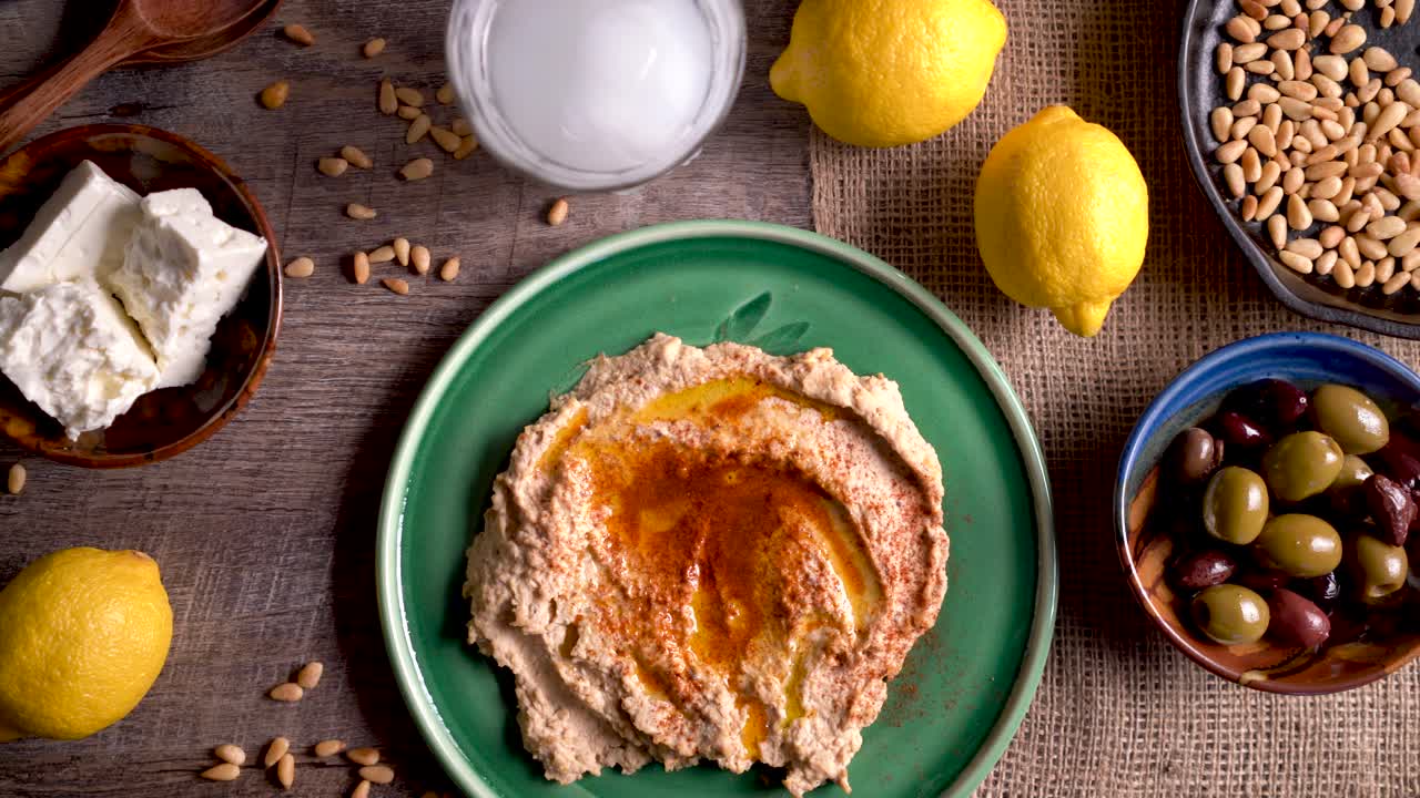 Ampia carrellata verso destra che mostra un piatto dall'alto di hummus con feta, olive, un bicchiere di ouzo, limoni, pinoli su un tavolo di legno rustico.